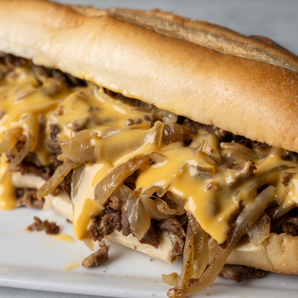 Close-up of a cheesesteak sandwich with melted cheese, steak, and caramelized onions on a white plate.